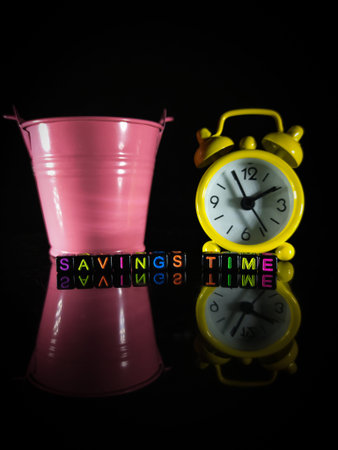 Selective focus.Word SAVINGS TIME with bucket,clock and reflection on a black background.Shot were...