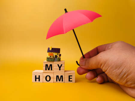 Hand holding red umbrella,miniature house and wooden cube with word MY HOME on a yellow background. Business concept.の写真素材