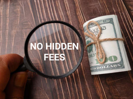 Hand holding magnifying glass and banknotes with text NO HIDDEN FEES on wooden background.の写真素材