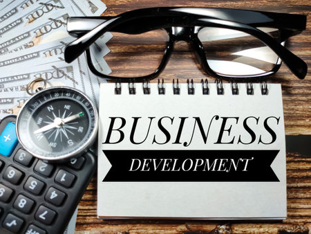 Business concept.Text BUSINESS DEVELOPMENT with compass,glasses,banknote and blur calculator on a brown wooden background.の写真素材
