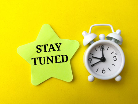 Top view colored sticky note and clock with text STAY TUNED on yellow background.