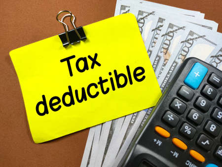 Business concept.Text Tax deductible on sticky note with calculator and banknote on brown background.の写真素材