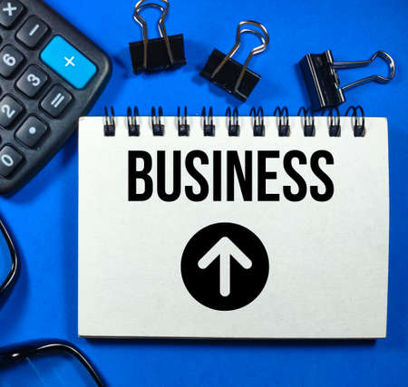 Business concept.Text Business and arrow icon on notebook with calculator,glasses and paper clips on blue background.の写真素材