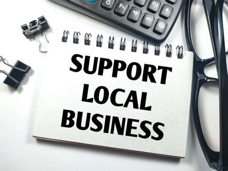 Business concept.Text SUPPORT LOCAL BUSINESS on notebook with paper clips,calculator and glasses on white background.の写真素材