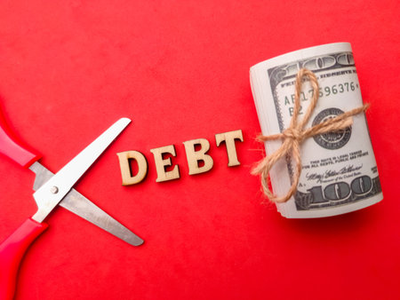Scissors, banknotes and wooden word with text DEBT on a red background.
