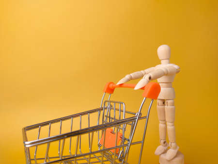 Wooden mannequin holding shopping trolley on a yellow background.の写真素材