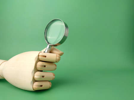 Wooden hand holding magnifying glass on a green background with copy space.の写真素材