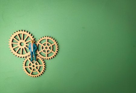 The image depicts a miniature figurine of a person in a suit standing on three wooden gears, arranged in a meshing configuration, all set against a plain green background.の写真素材