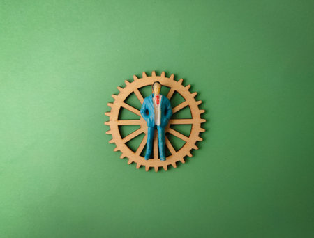 The image depicts a small figurine of a businessman standing on a wooden gear wheel.の写真素材