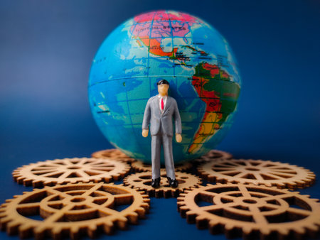 The image depicts a miniature businessman figure standing in front of a globe, with several wooden gears surrounding the base of the globe.の写真素材