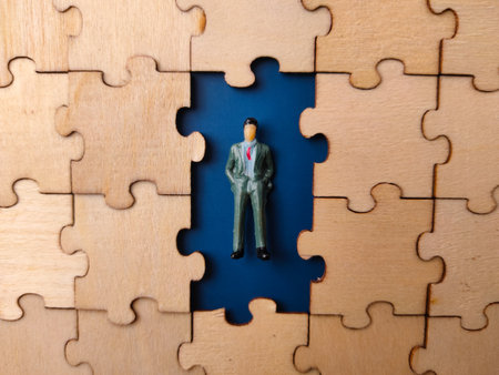 The image depicts a miniature figure of a businessman standing in a puzzle piece-shaped hole, surrounded by completed wooden puzzle pieces.の写真素材
