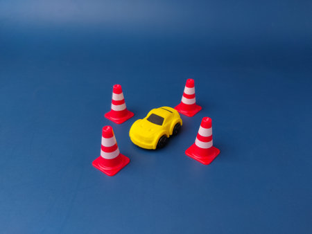 This image shows a small yellow toy car surrounded by four miniature traffic cones on a blue background.の写真素材