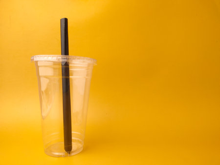 The image shows a clear disposable plastic cup with a lid and a black straw on a yellow background.の写真素材