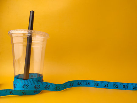 The image shows a clear disposable plastic cup with a black straw, encircled by a blue measuring tape on yellow background.の写真素材