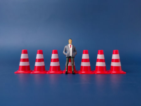 The image shows a small figurine of a man in a business suit standing among a line of red and white traffic cones.の写真素材