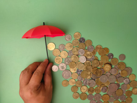 The hand holding a small red umbrella over a pile of coins symbolizes the act of safeguarding one's finances from potential losses or risks.の写真素材