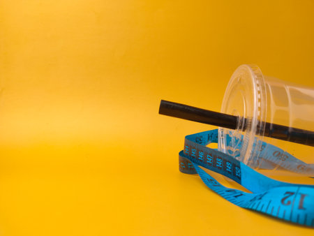 The image shows a clear disposable plastic cup with a black straw, encircled by a blue measuring tape on yellow background.の写真素材