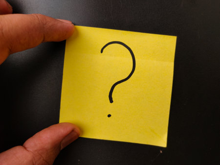 The image shows a hand holding a yellow sticky note with a question mark drawn on it.の写真素材
