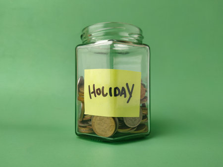 The image shows a glass jar filled with coins and a yellow sticky note on it with the word Holiday written in black marker.の写真素材