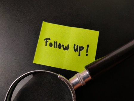 The image shows a yellow sticky note with the words Follow up! written on it, with a magnifying glass in the foreground.の写真素材
