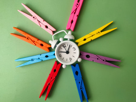 White alarm clock surrounded by several colorful wooden clothes pegs on a green background.