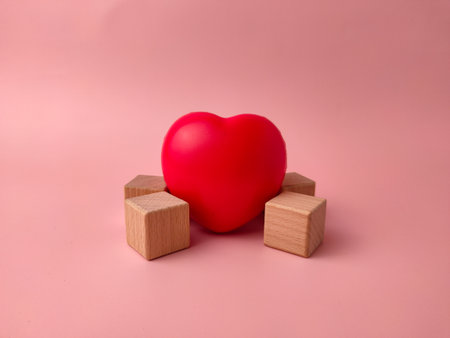 Red heart-shaped object positioned between two small wooden blocks on a pink background.の写真素材