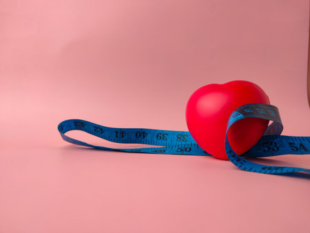 The image features a red heart object encircled by a blue tape measure, set against a pink background.の写真素材