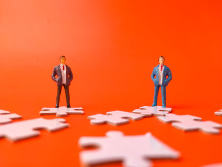 The image depicts miniature figurines of two businessmen standing on separate white jigsaw puzzle pieces against a bright orange background.の写真素材