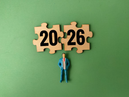 The image depicts two jigsaw puzzle pieces forming the number 2026, with a miniature figure of a businessman standing below them on a green background.の写真素材