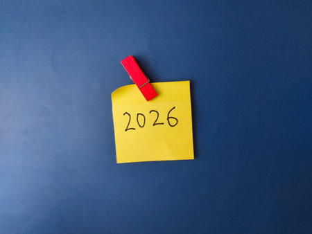 The image shows a yellow sticky note with the year 2026 written in black ink, pinned to a blue...