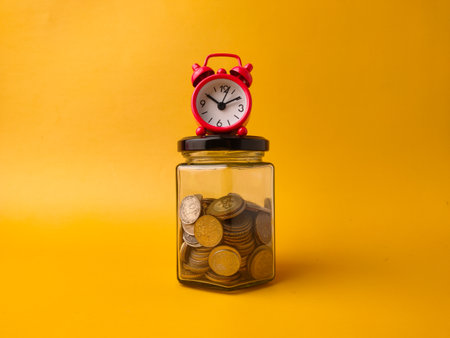 The image shows a red alarm clock sitting on top of a glass jar filled with coins.の写真素材
