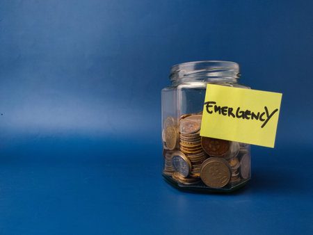 The image displays a glass jar filled with coins, featuring a yellow sticky note with the word Emergency written on it.の写真素材