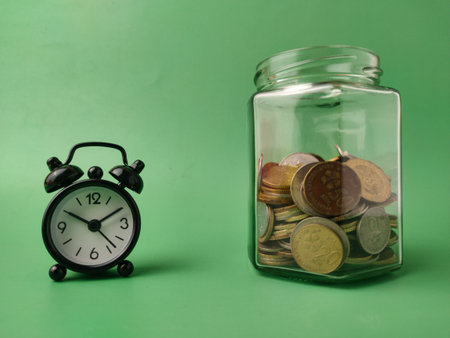 The image displays a clear glass jar filled with coins and a small, black alarm clock on a green background.の写真素材