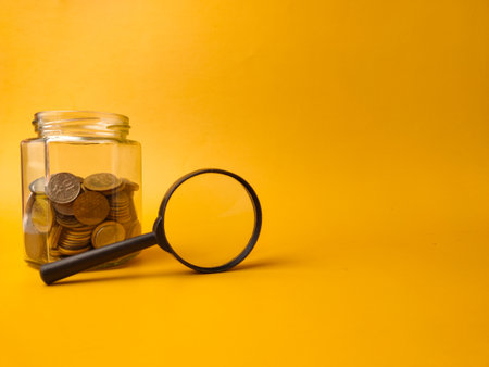 The image displays a clear glass jar filled with coins and a magnifying glass resting against it, set against a yellow background.の写真素材