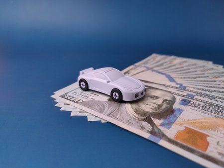 The image displays a toy car on a pile of US one-hundred-dollar bills, representing concepts such as car insurance, saving money, or purchasing a vehicle.の写真素材