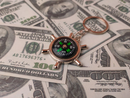 small metal compass keychainÂ resting on a bed of US hundred-dollar bills.