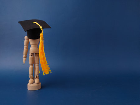 The image features a wooden artist's mannequin wearing a miniature graduation cap and yellow tassel on a blue background.の写真素材