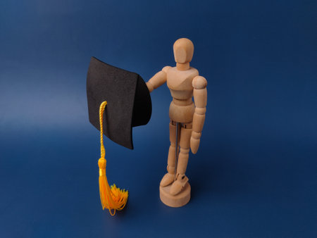 Wooden artist's mannequin holding a miniature square academic cap, commonly known as a mortarboard, on a blue background.の写真素材