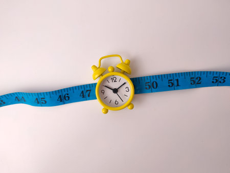 Yellow alarm clock positioned on a blue measuring tape on a white background.の写真素材
