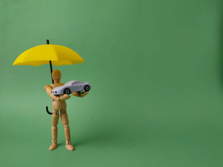 The wooden figure holding a yellow umbrella over the toy car symbolizes an insurance agent or coverage providing safety.の写真素材