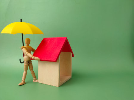 Wooden artist's manikin standing inside a wooden house model with a red roof, topped by a small yellow umbrella.の写真素材