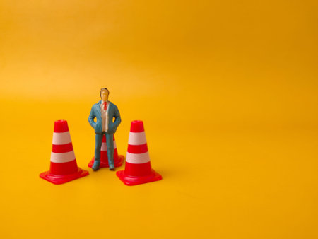 Miniature businessman figurine positioned between traffic cones on a yellow background.