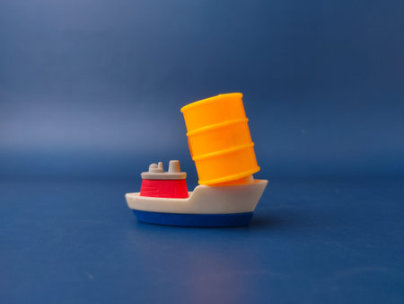 Conceptual image of a miniature toy ship carrying an oversized yellow plastic barrel against a solid...