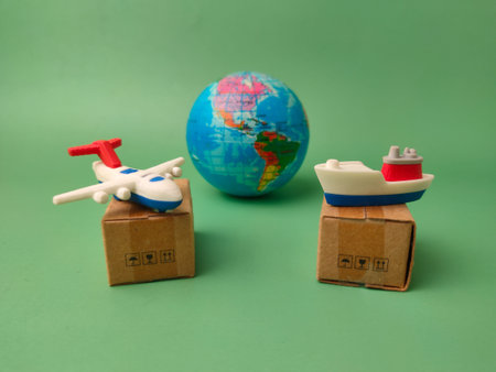 Small toy airplane and a cargo ship, each resting on a miniature cardboard shipping box, with a...