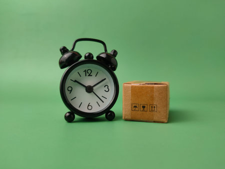 Small black alarm clock and a single miniature cardboard shipping box on a green background.