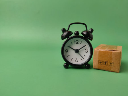 Small black alarm clock and a single miniature cardboard shipping box on a green background.