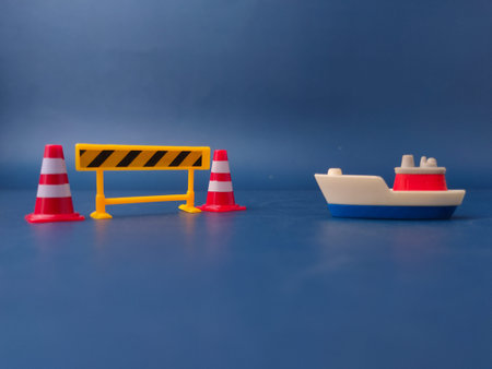 A toy ship facing a yellow construction barrier and traffic cones on a blue background.の写真素材