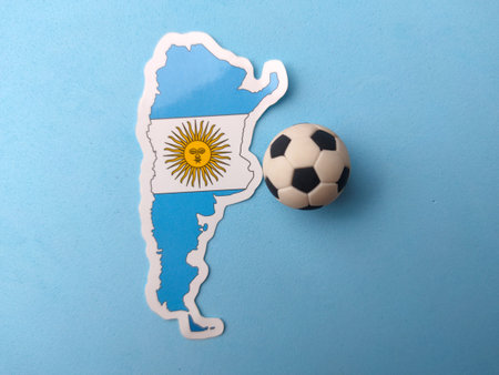 An Argentina map sticker displaying its national flag is positioned next to a miniature football on a blue background.
