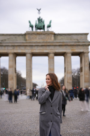 A woman in a stylish grey coat stands before the iconic Berlin gateの写真素材