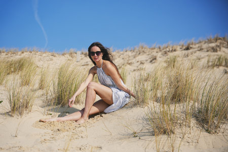 Barefoot woman in white dress relaxing on dune with tall grass under blue skyの写真素材